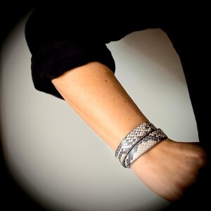 Peut inclure: Un bracelet à motif peau de serpent enroulé autour d'un poignet. Le bracelet présente un motif répété d'écailles grises et blanches. Le bras est partiellement visible, avec une manche noire en arrière-plan.
