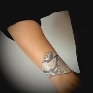 Può includere: Un bracciale a polsino in pelle con motivo a pelle di serpente. Il bracciale è beige, grigio e nero, con una forma curva. Il bracciale è indossato sull'avambraccio.