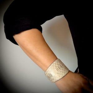 May include: A wide, textured cuff bracelet in a light, neutral color. The bracelet appears to be made of a material with a scale-like pattern. The cuff is worn on a person's arm, with a black sleeve in the background.