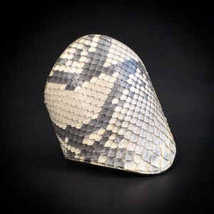 Peut inclure: Un accessoire à motif peau de serpent, possiblement un chapeau ou un bracelet, sur fond noir. L'objet présente une texture en écailles avec une palette de gris, de blanc et de noir. Le design suggère une esthétique naturelle, inspirée des reptiles.