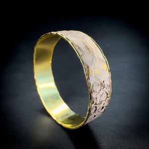 Könnte beinhalten: Ein goldfarbenes Armreif-Armband mit einem strukturierten Band mit floralem Muster in Weiß und Gold. Die Innenseite und die Kanten des Armbands sind goldfarben, was einen Kontrast erzeugt. Das Armband wird vor dunklem Hintergrund gezeigt.