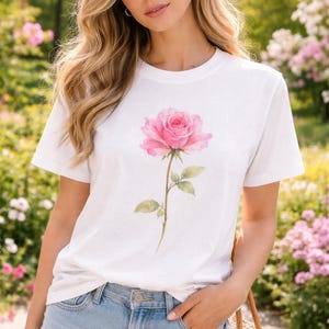 May include: White t-shirt featuring a large pink rose illustration. The rose has detailed petals and a green stem with leaves. The shirt is paired with light blue denim shorts. The background shows a garden with pink flowers.