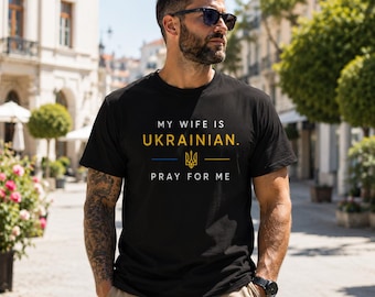 May include: A black short-sleeved t-shirt featuring the white text MY WIFE IS, the yellow text UKRAINIAN, a blue and yellow horizontal line with a gold trident symbol in the center, and the white text PRAY FOR ME printed on the front chest.