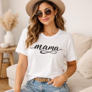 Op de afbeelding: Wit T-shirt met het woord "mama" in zwart script. Het woord is gestileerd met een doorlopend lijnontwerp. De persoon draagt een zonnebril, hoed en riem.