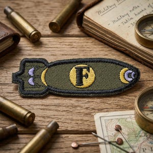 May include: An olive green embroidered patch in the shape of a bomb, featuring a yellow circle with a black "F" and two smaller yellow and purple accents. The patch is outlined in black and sits on a wooden surface with other military-themed items.