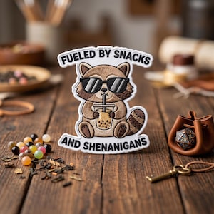 May include: Embroidered patch featuring a raccoon wearing sunglasses and holding a boba drink. The patch has the text "Fueled by Snacks" and "And Shenanigans" in a white, curved font. The raccoon is brown and tan.