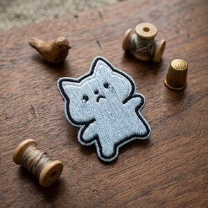 May include: A light blue embroidered cat patch with a black outline, featuring a sad face. The patch is surrounded by a wooden bird figurine, a gold thimble, and spools of thread on a wooden surface.