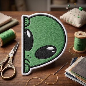 May include: A green alien patch with black eyes and a white border. The patch is on a wooden surface with sewing supplies, including scissors, thread, and a pin cushion. The patch is a fun, quirky design.