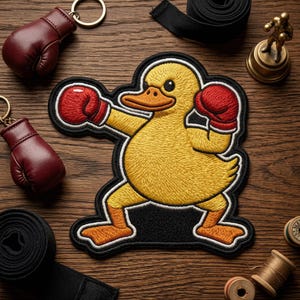 May include: Embroidered yellow duck patch wearing red boxing gloves, with a black border. Two miniature red boxing glove keychains and a small gold statue are also visible on a wooden surface.