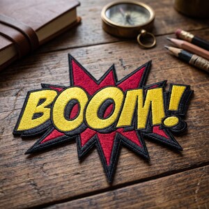 May include: Embroidered patch with the word "BOOM!" in yellow, outlined in black, set against a red burst design. The patch is on a wooden surface, with a notebook, compass, and pencils in the background.