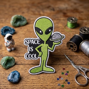 May include: Embroidered patch featuring a green alien holding a planet, with the text "SPACE IS COOL." The patch is surrounded by sewing supplies, including spools of thread, scissors, and decorative gems. The alien has black eyes and is outlined in black.