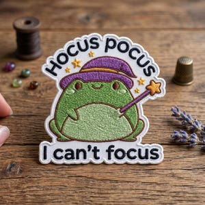 May include: Embroidered patch featuring a green frog wearing a purple wizard hat and holding a star wand. The patch reads "Hocus Pocus" above the frog and "I can't focus" below. The patch is on a wooden surface.