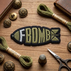 May include: An embroidered patch in the shape of a bomb with the word "FBOMB" in white and olive green. The patch is surrounded by black stitching. The image also includes spools of green thread, brass buttons, a thimble, and scissors, all arranged on a wooden surface.