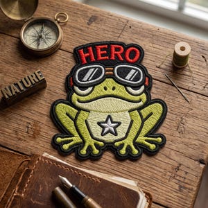 May include: Embroidered patch featuring a cartoon frog wearing goggles and a "HERO" banner. The frog is green and yellow with a white star on its chest. The patch is on a wooden surface with a compass and the word "NATURE".