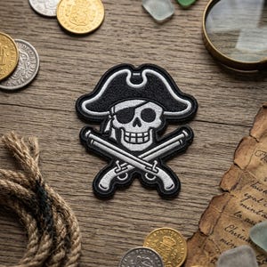 May include: Embroidered pirate patch featuring a skull wearing a tricorn hat and an eye patch, with crossed pistols below. The patch is black and white, with detailed stitching. The design is set against a wooden background with coins and a rope.