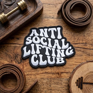 May include: Black and white embroidered patch with the text "ANTI SOCIAL LIFTING CLUB". The patch is on a wooden surface with a small dumbbell and leather straps. The patch has a black background and white lettering.