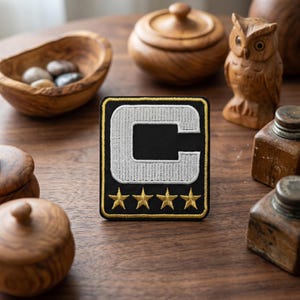 May include: A black and gold embroidered patch featuring a white letter "C" and four gold stars. The patch has a black background with a gold border. The patch is likely a sports team captain's insignia or a similar emblem.