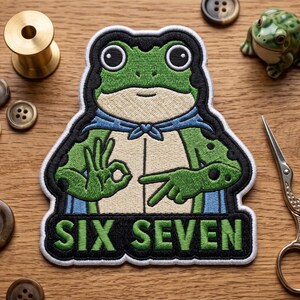 May include: Embroidered patch featuring a green frog with a blue cape, making a hand gesture. The patch has the words "SIX SEVEN" in a black banner. The frog is surrounded by a black border. Other sewing supplies are in the background.