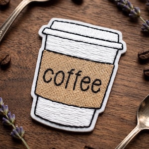 May include: Embroidered patch in the shape of a coffee cup with the word "coffee" in black script on a tan burlap-like wrap. The cup is white with a white lid. The patch is on a wooden surface with coffee beans and lavender.