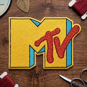 May include: Embroidered yellow MTV logo patch with blue and red accents. The patch is rectangular with the letters "MTV" in a bold, stylized font. A gold guitar pick with a lightning bolt design is also visible.