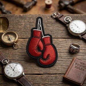 May include: A red embroidered boxing gloves patch with a black border. The patch is on a wooden surface with a compass, stopwatch, ring, and a small book titled "OLD BOXING RULES".