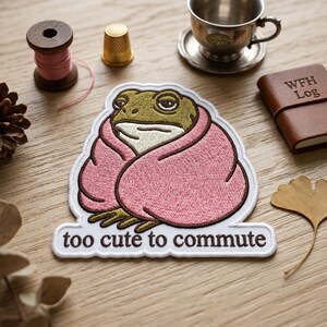 May include: Embroidered patch featuring a cartoon frog wrapped in a pink blanket. The frog has a green head and is embroidered with a white border. The text "too cute to commute" is below the frog.
