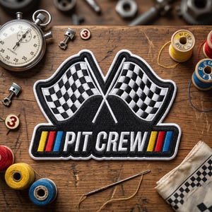 May include: A black and white checkered flag patch with the words "PIT CREW" in white. The patch has red, yellow, and blue stripes. A stopwatch, spools of thread, and other tools are arranged on a wooden surface.