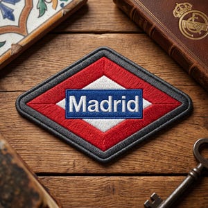 May include: Embroidered patch featuring the Madrid Metro logo. The patch has a red and white diamond shape with a blue rectangle in the center, displaying the city's name in white. The patch is bordered by a gray outline.