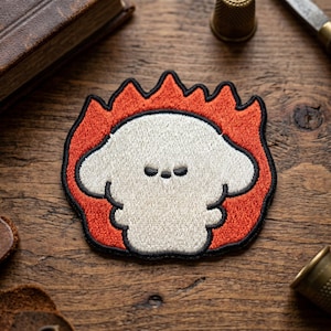 May include: Embroidered patch featuring a cartoon dog-like character in a fiery design. The patch has a white face with three small black dots for eyes, surrounded by orange flames with a black outline. The patch is on a wooden surface.