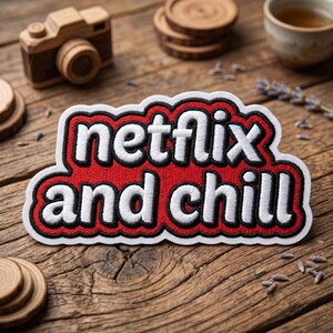 May include: A red, white, and black embroidered patch with the words "Netflix and Chill". The patch is surrounded by a white border and a black outline. The patch is on a wooden surface, with a wooden camera and coasters in the background.