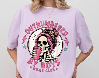 May include: Orchid-colored t-shirt with a graphic featuring a skeleton with pink hair and a leopard print headband, holding a pink cup. The text reads "OUTNUMBERED BY BOYS * MOMS CLUB *" in a circular design.