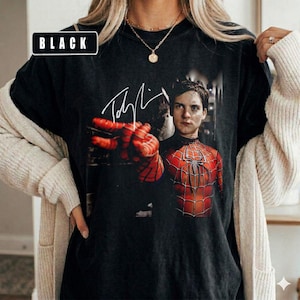 Puede incluir: Camiseta negra con un gr&aacute;fico de Spider-Man y una firma. El dise&ntilde;o muestra al superh&eacute;roe con su traje rojo y negro, con un primer plano de su mano. La palabra "BLACK" est&aacute; en un rect&aacute;ngulo blanco en la esquina superior izquierda.