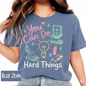 May include: A blue jean colored t-shirt featuring the pink and white text "You Can Do Hard Things" with a yellow lightbulb graphic, a 3D printer, a microscope, a laptop, a water bottle, and art supplies printed in various colors on the front.