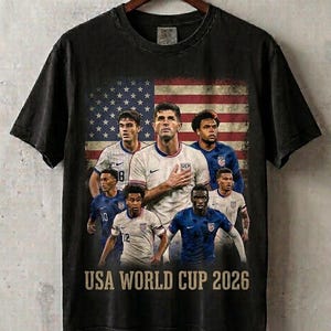 May include: A black short-sleeve t-shirt featuring a graphic print of six soccer players in team uniforms positioned in front of a faded American flag. The text USA WORLD CUP 2026 is printed in a tan, distressed font at the bottom of the graphic.