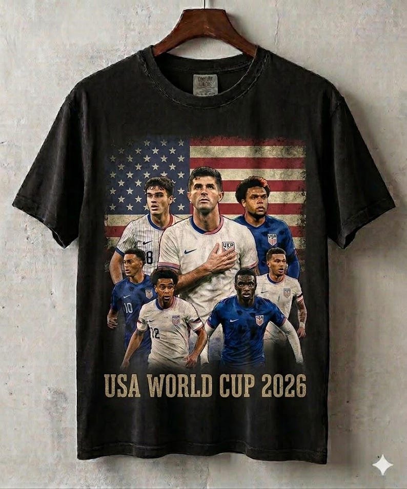 May include: A black short-sleeve t-shirt featuring a graphic print of six soccer players in team uniforms positioned in front of a faded American flag. The text USA WORLD CUP 2026 is printed in a tan, distressed font at the bottom of the graphic.