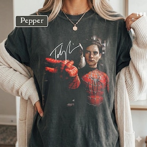Puede incluir: Camiseta gris oscuro con un gr&aacute;fico de Spider-Man y una firma. La imagen muestra a Spider-Man con su traje rojo y negro, con una mano extendida. La palabra "Pepper" est&aacute; en un rect&aacute;ngulo blanco en la esquina superior izquierda.