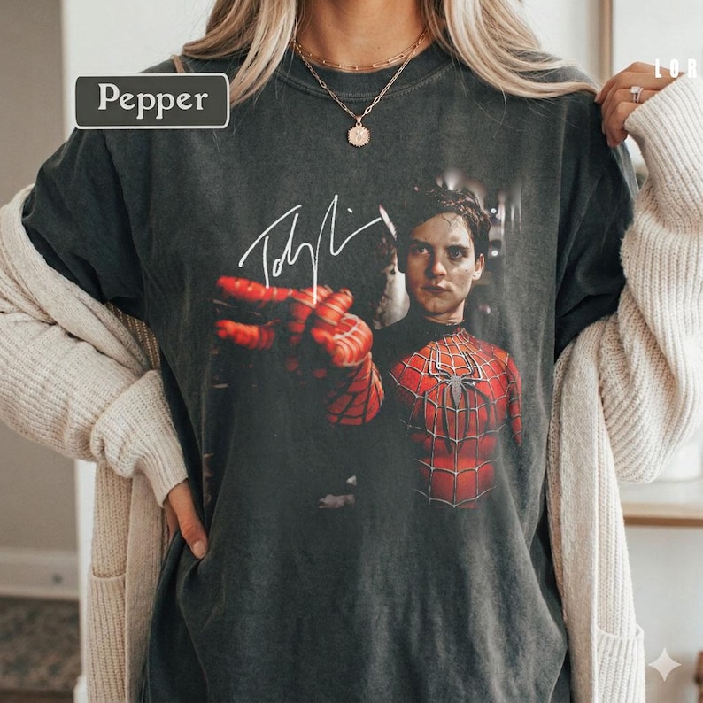 Puede incluir: Camiseta gris oscuro con un gr&aacute;fico de Spider-Man y una firma. La imagen muestra a Spider-Man con su traje rojo y negro, con una mano extendida. La palabra "Pepper" est&aacute; en un rect&aacute;ngulo blanco en la esquina superior izquierda.