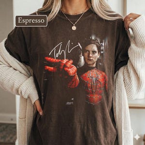 Puede incluir: Camiseta color espresso con un gr&aacute;fico de Spider-Man y una firma. La imagen muestra al personaje con su traje rojo y negro, con un primer plano de su mano. La camiseta la lleva una persona.