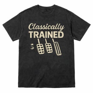 May include: Dark gray t-shirt with the words "Classically TRAINED" in a distressed font. Below the text are illustrations of a clutch, brake, and accelerator pedals, and a coffee cup.