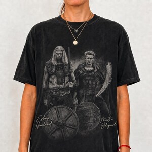 May include: A black short-sleeve t-shirt featuring a graphic print of two figures in Viking-style attire. One figure holds a sword and the other a drinking horn, with round shields below. The text Erling Haaland and Martin Odegaard is printed.