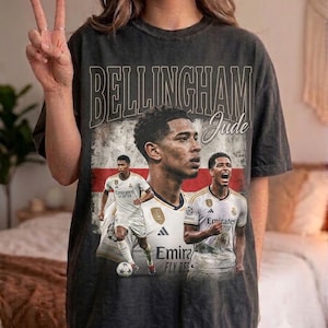 May include: A charcoal gray short-sleeve t-shirt featuring a vintage-style graphic of soccer player Jude Bellingham. The design includes the text "BELLINGHAM" and "Jude" over a faded English flag background with multiple images of the athlete.
