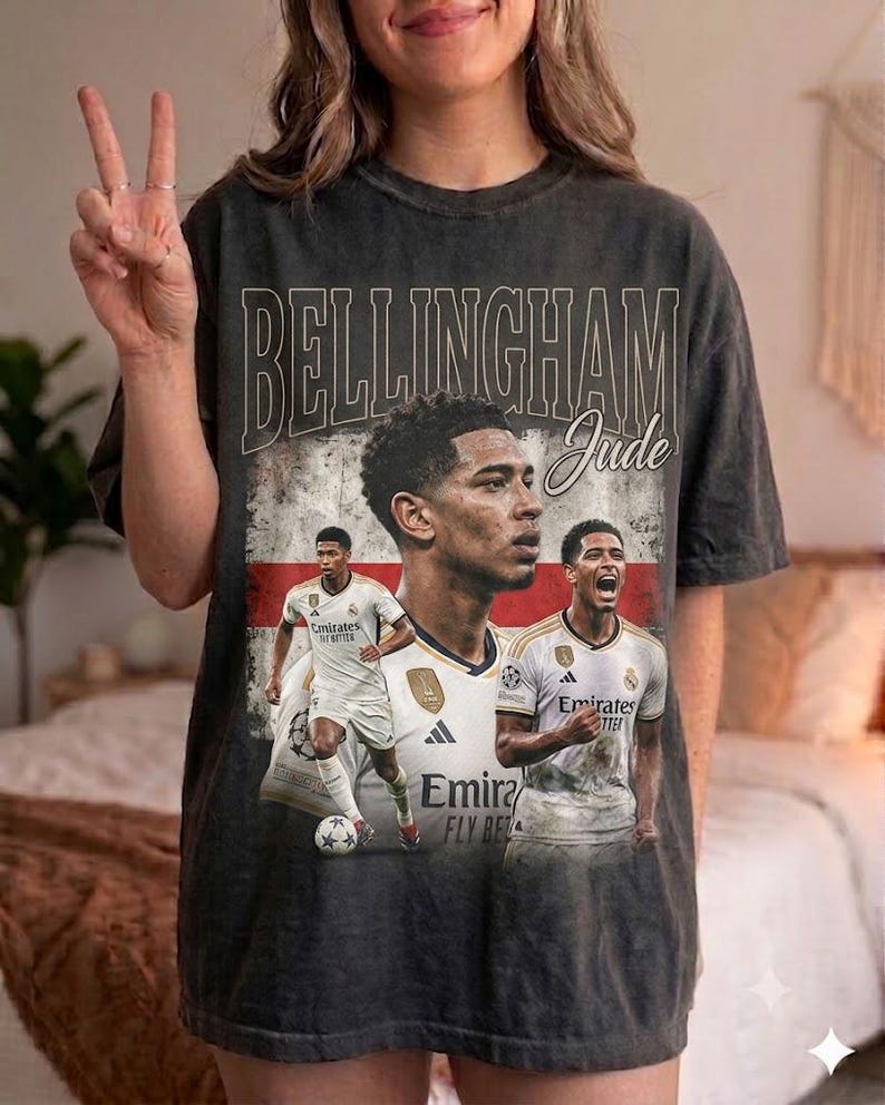 May include: A charcoal gray short-sleeve t-shirt featuring a vintage-style graphic of soccer player Jude Bellingham. The design includes the text "BELLINGHAM" and "Jude" over a faded English flag background with multiple images of the athlete.