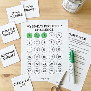 Könnte beinhalten: Ein 30-Tage-Entrümpelungs-Challenge mit nummerierten Kreisen, einem Glas mit Etiketten und Karten für verschiedene Bereiche. Ein grüner Marker und eine Schere sind ebenfalls vorhanden. Der Kalender ist mit grünen Kreisen markiert.