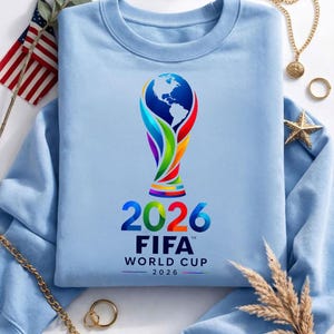 May include: A light blue crewneck sweatshirt featuring a colorful graphic of the FIFA World Cup 2026 trophy, which includes a globe design. Below the trophy, the text reads 2026 FIFA WORLD CUP 2026 in bold, dark blue and multicolored lettering.