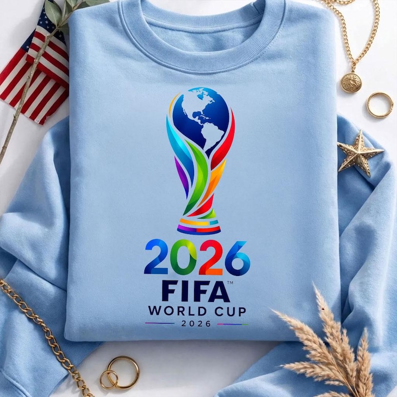 May include: A light blue crewneck sweatshirt featuring a colorful graphic of the FIFA World Cup 2026 trophy, which includes a globe design. Below the trophy, the text reads 2026 FIFA WORLD CUP 2026 in bold, dark blue and multicolored lettering.