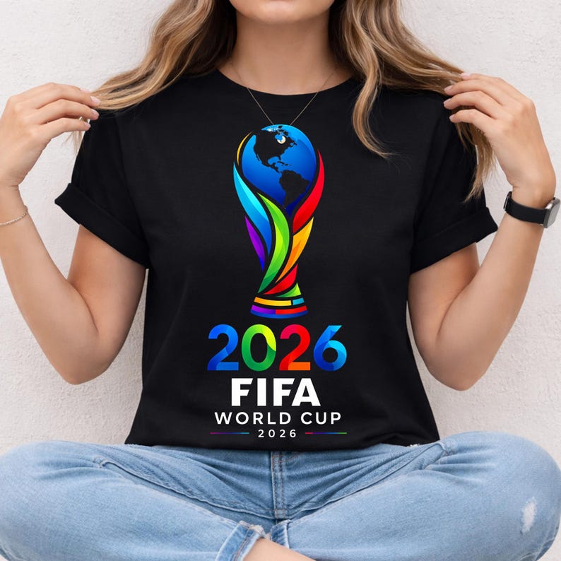 May include: A black short-sleeve t-shirt featuring a colorful graphic of the FIFA World Cup 2026 trophy with a globe at the top. Below the trophy, the text reads 2026 FIFA WORLD CUP 2026 in bold, multi-colored and white lettering.