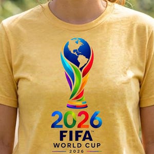 May include: A heathered yellow short-sleeve t-shirt featuring the FIFA World Cup 2026 logo. The graphic displays a stylized trophy shape with a blue globe at the top, colorful ribbons, and the text 2026 FIFA WORLD CUP 2026 printed in blue and red.