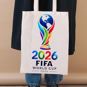 May include: A white canvas tote bag featuring a colorful graphic of the FIFA World Cup 2026 trophy. The design includes a stylized globe at the top with vibrant red, green, blue, and yellow accents, followed by the text 2026 FIFA WORLD CUP 2026.