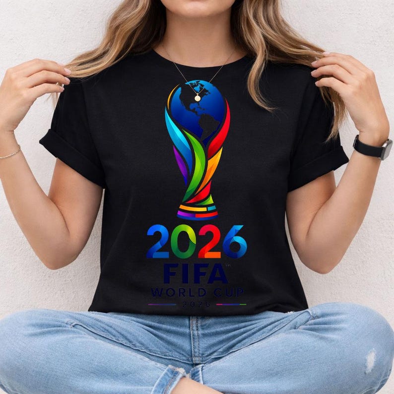 May include: A black t-shirt featuring a colorful graphic of the 2026 FIFA World Cup trophy. The design includes a stylized globe at the top, vibrant red, green, and blue accents, and the text 2026 FIFA WORLD CUP printed in bold, multi-colored lettering.