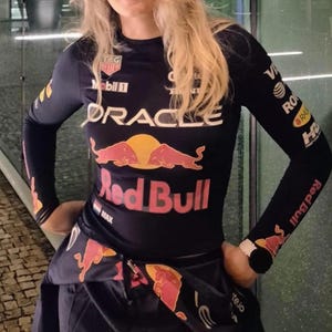 Puede incluir: Camiseta negra de manga larga con el logo de Red Bull y la marca Oracle. La camiseta presenta un gráfico de sol y el texto "Red Bull" en rojo. Se ven logotipos y texto adicionales en las mangas.